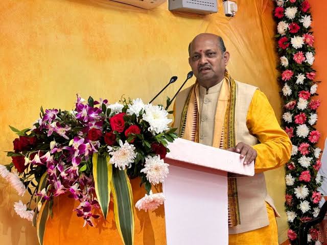 Manmohan Samal speaking at an indoor function from a lectern flanked by floral arrangements.