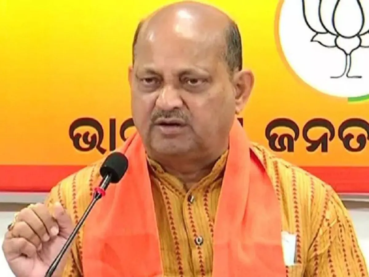 Manmohan Samal speaking into a microphone at a party press briefing, before a backdrop bearing the party name in Odia.