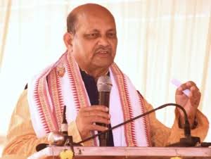 Manmohan Samal addressing a gathering from a lectern, wearing a striped Odisha shawl.