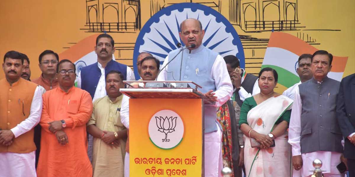 Manmohan Samal speaking from a lectern bearing the Bharatiya Janata Party Odisha emblem, with party colleagues standing behind him in front of a large tricolour backdrop.