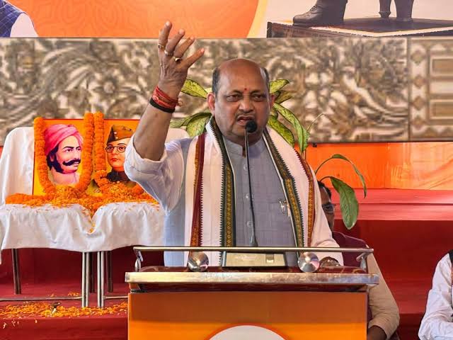 Manmohan Samal speaking from a lectern with one hand raised, beside garlanded portraits displayed at a commemorative gathering.