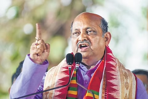 Manmohan Samal addressing an outdoor gathering with an index finger raised, wearing a purple shirt and a striped Odisha shawl.