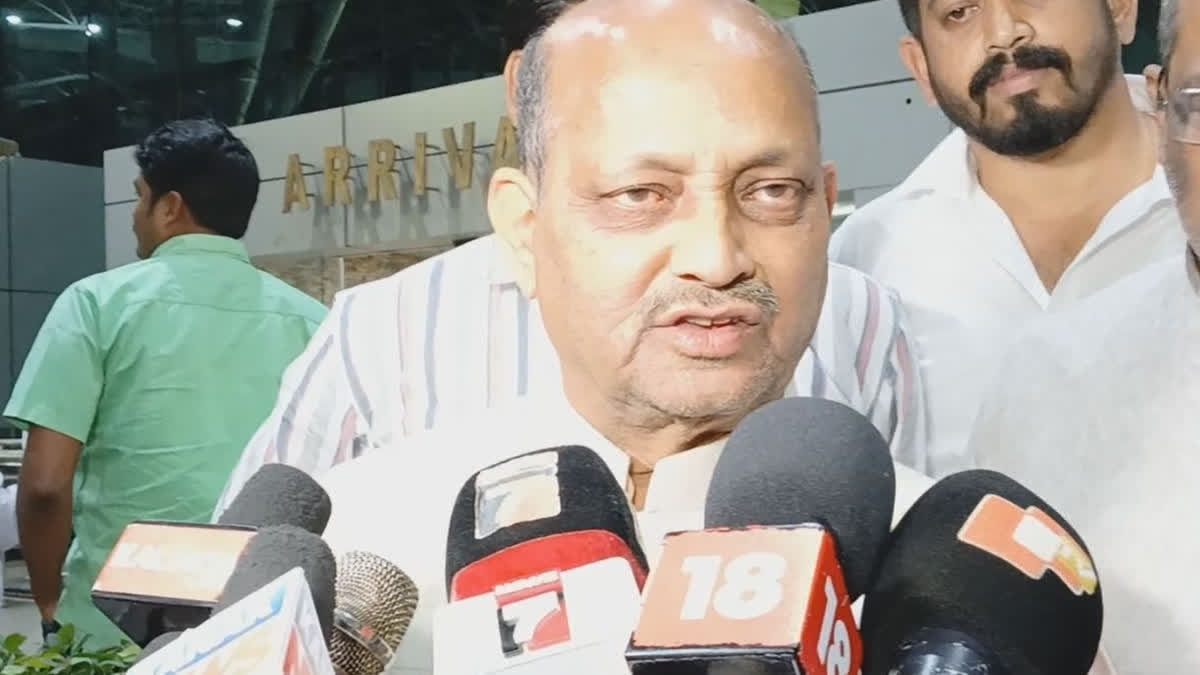 Manmohan Samal speaking to a bank of television microphones in an airport arrivals hall.